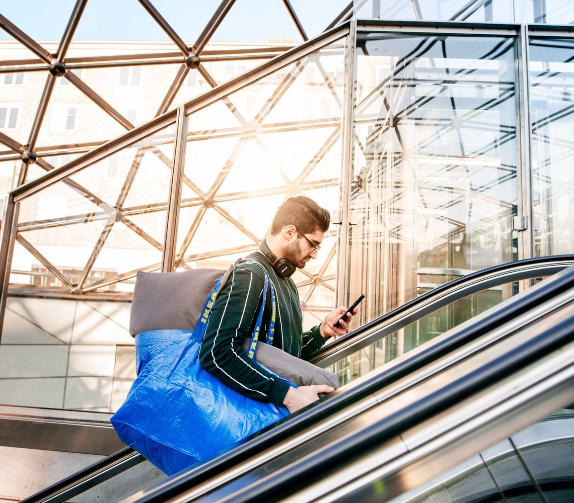 Man going up in an escalator with a blue IKEA bag over his shoulder