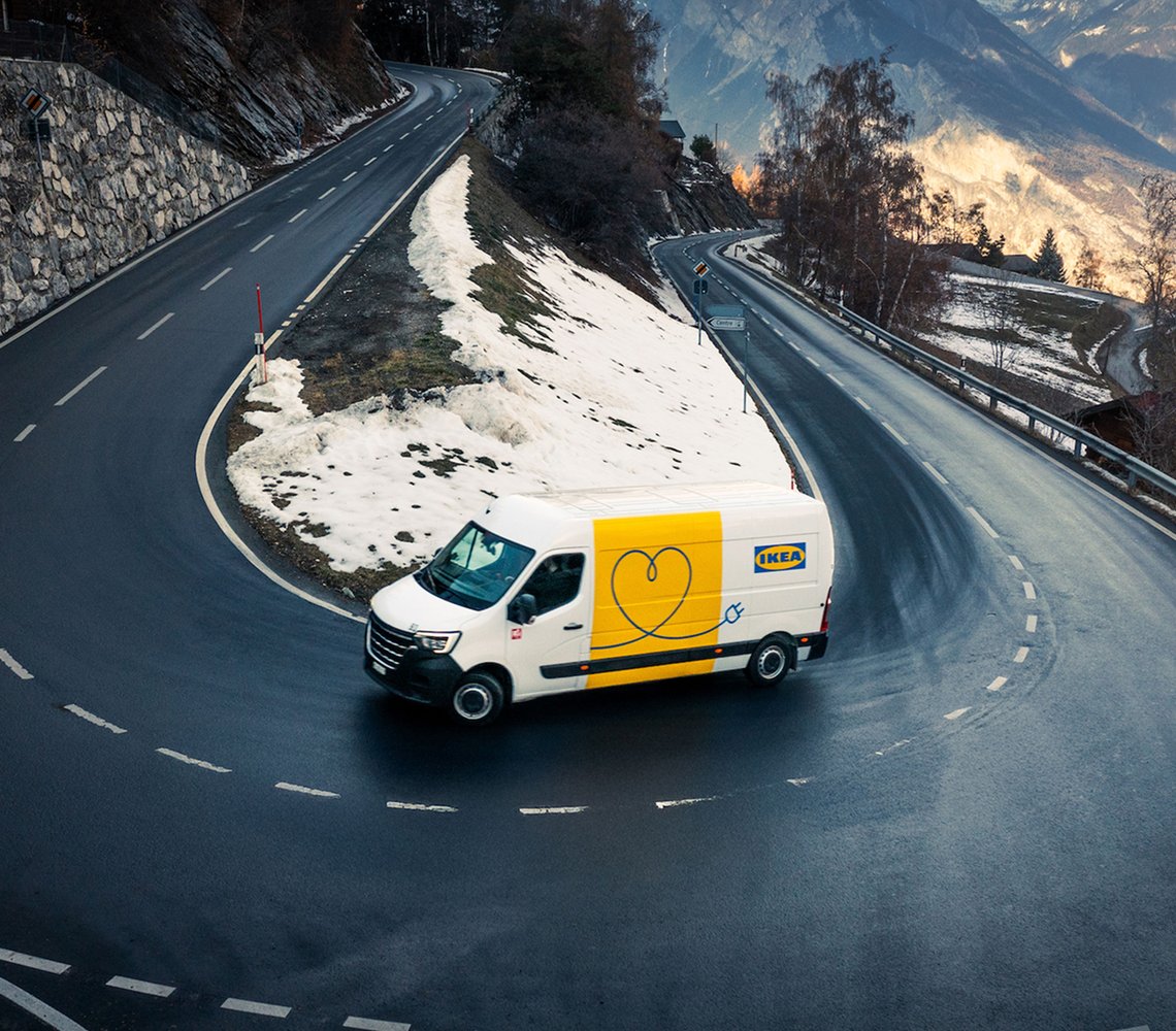 IKEA delivery van driving on a winding mountain road with snow patches and scenic valley views in the background.