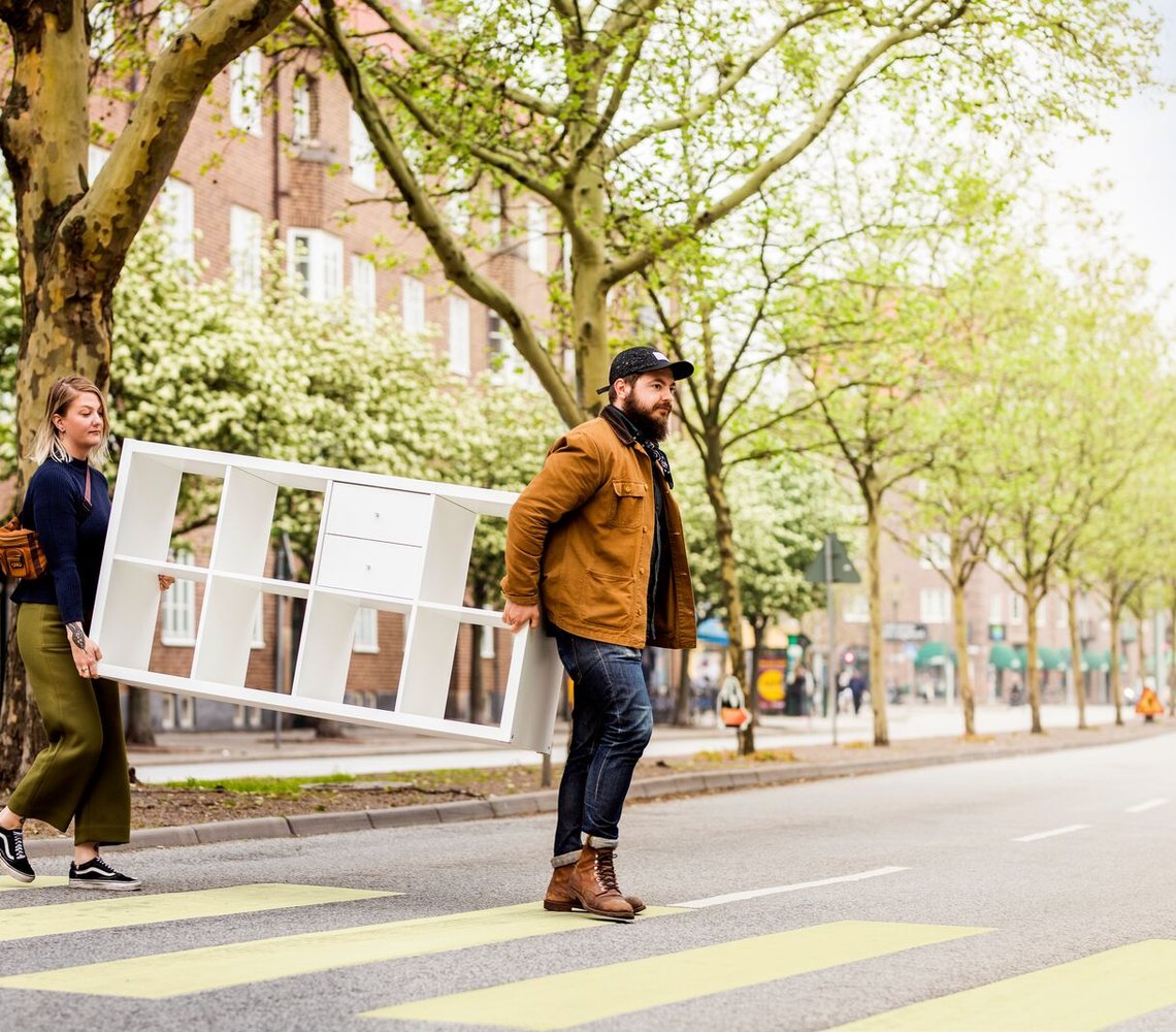 Two persons carrying a KALLAX over the street.