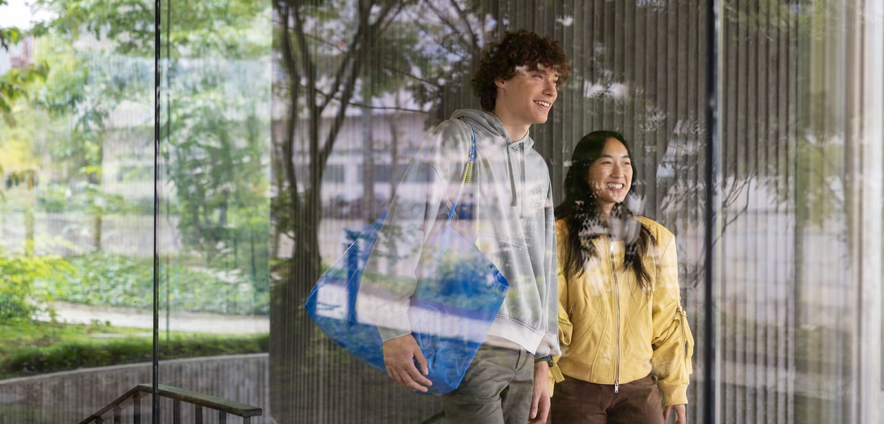 Two people walking behind a window with a blue FRAKTA bag.