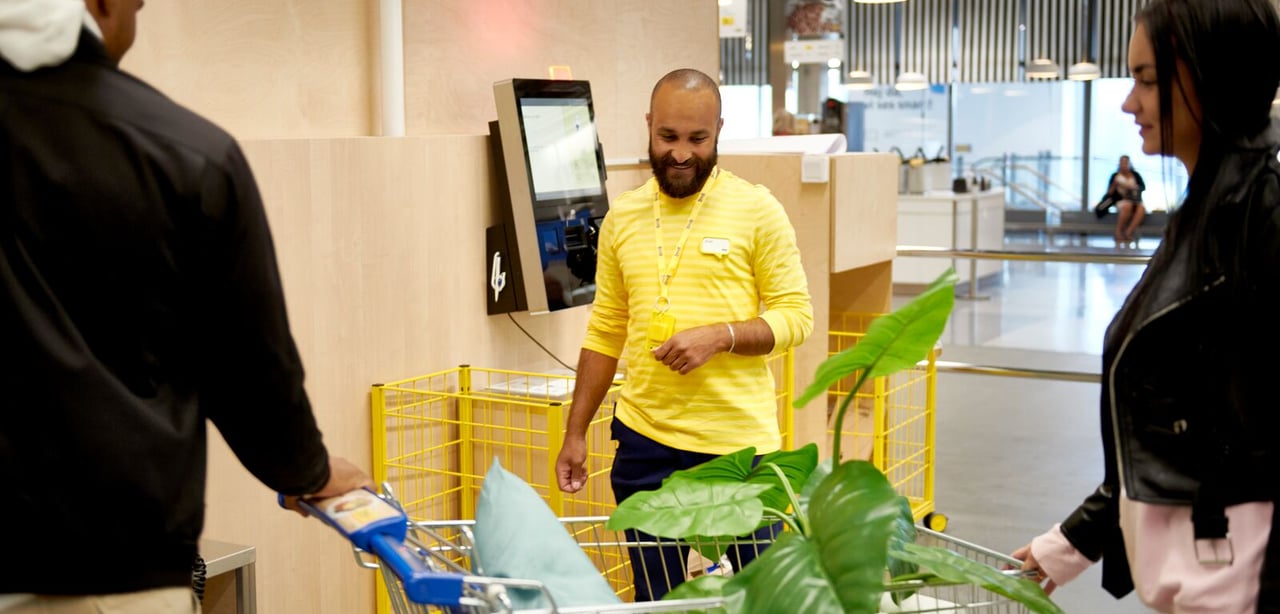 A smiling IKEA employee helping two customers in the self service check-out area.