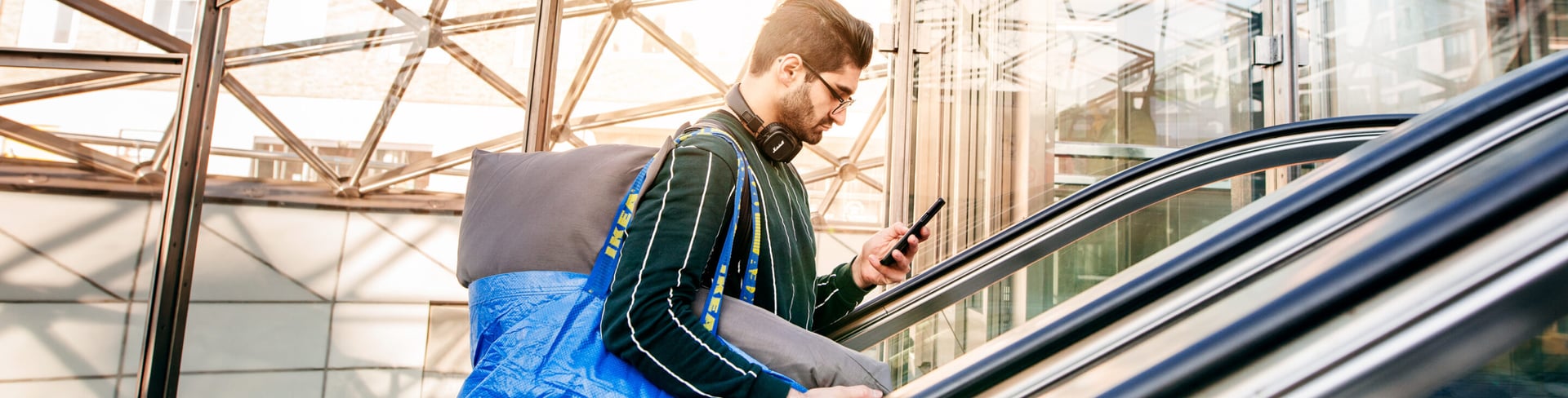 Young man carrying IKEA FRAKTA blue shopping bag and grey cushion on an escalator in a modern glass building, checking his phone after shopping at IKEA