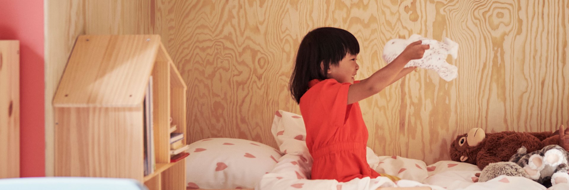 A child wearing a red dress sits on a bed playing with the soft toy SKOGSDUVA while smiling big.