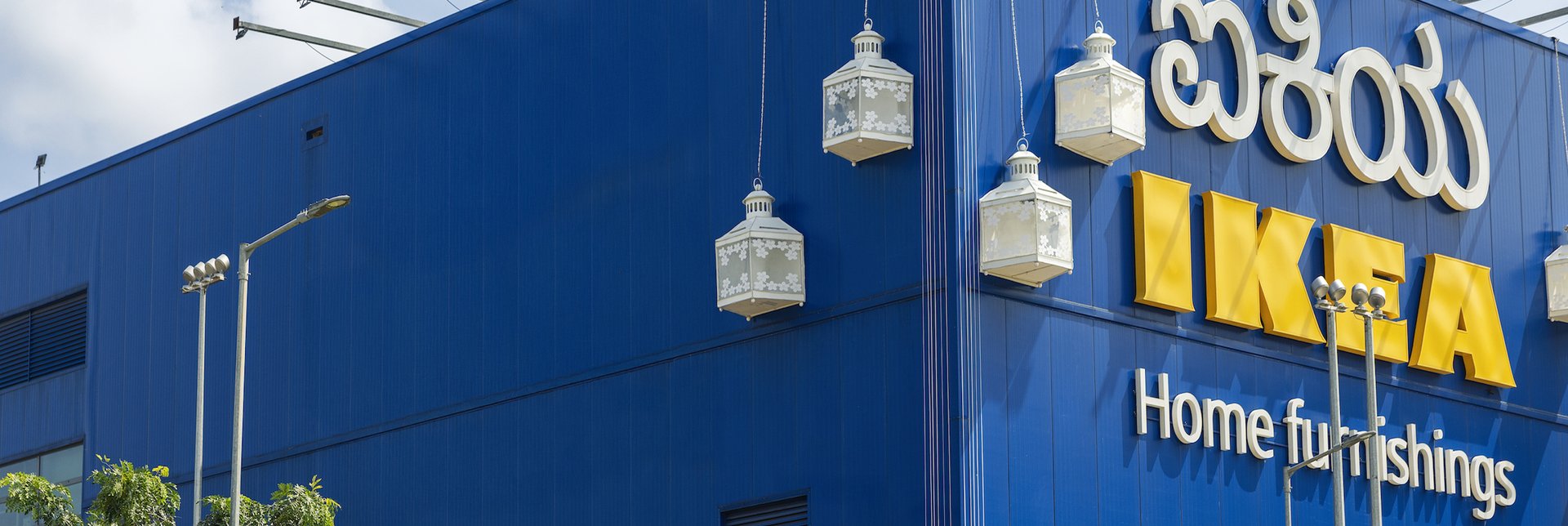 Exterior of an IKEA store with a blue facade, large yellow IKEA sign, and decorative white lanterns hanging on the building.