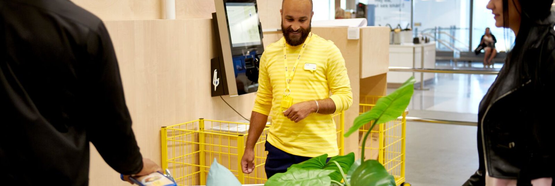 A smiling IKEA employee helping two customers in the self service check-out area.