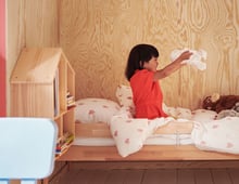 A child wearing a red dress sits on a bed playing with the soft toy SKOGSDUVA while smiling big.