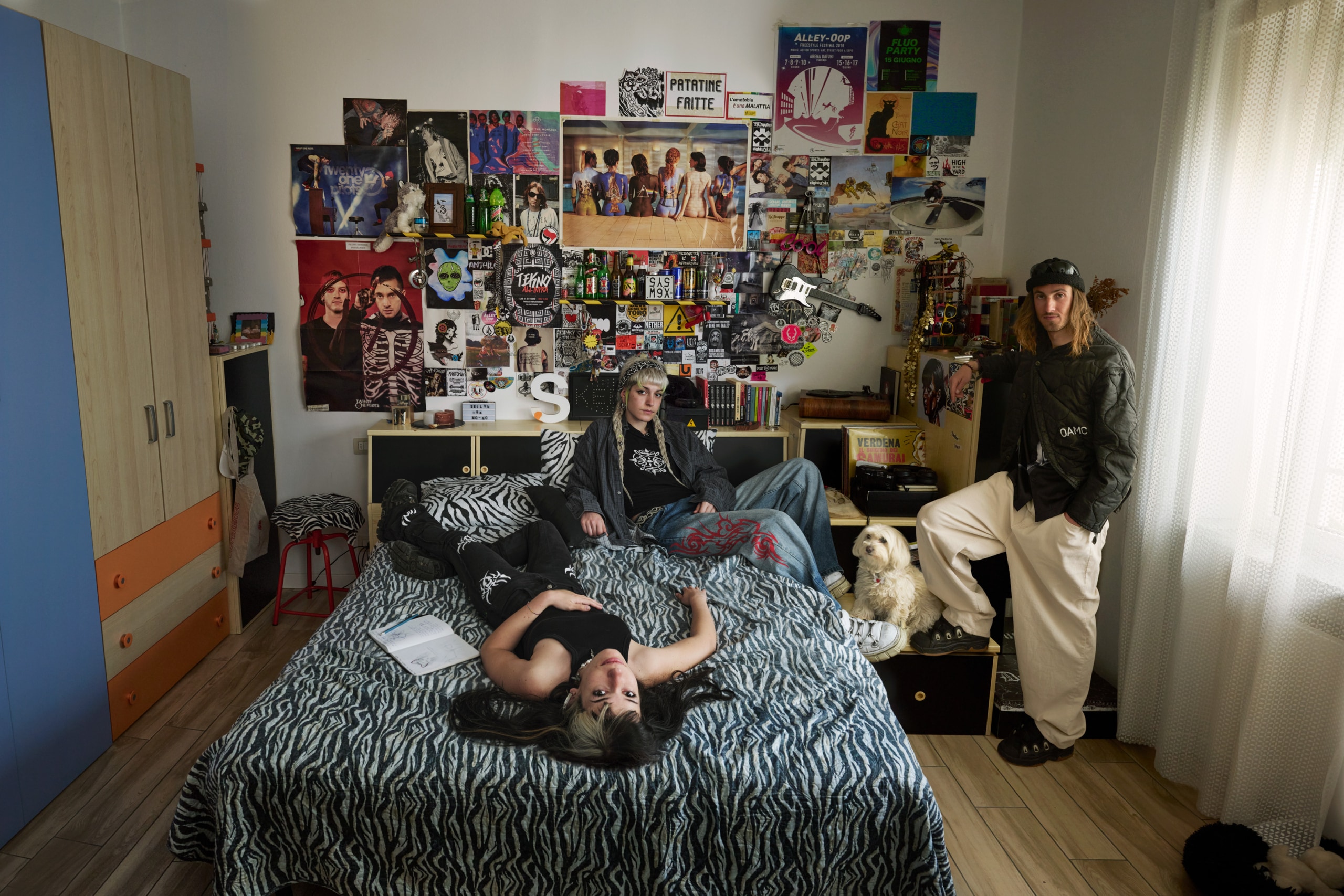 Silvia in her room with two friends, the wall behind her bed is covered with pictures and posters.