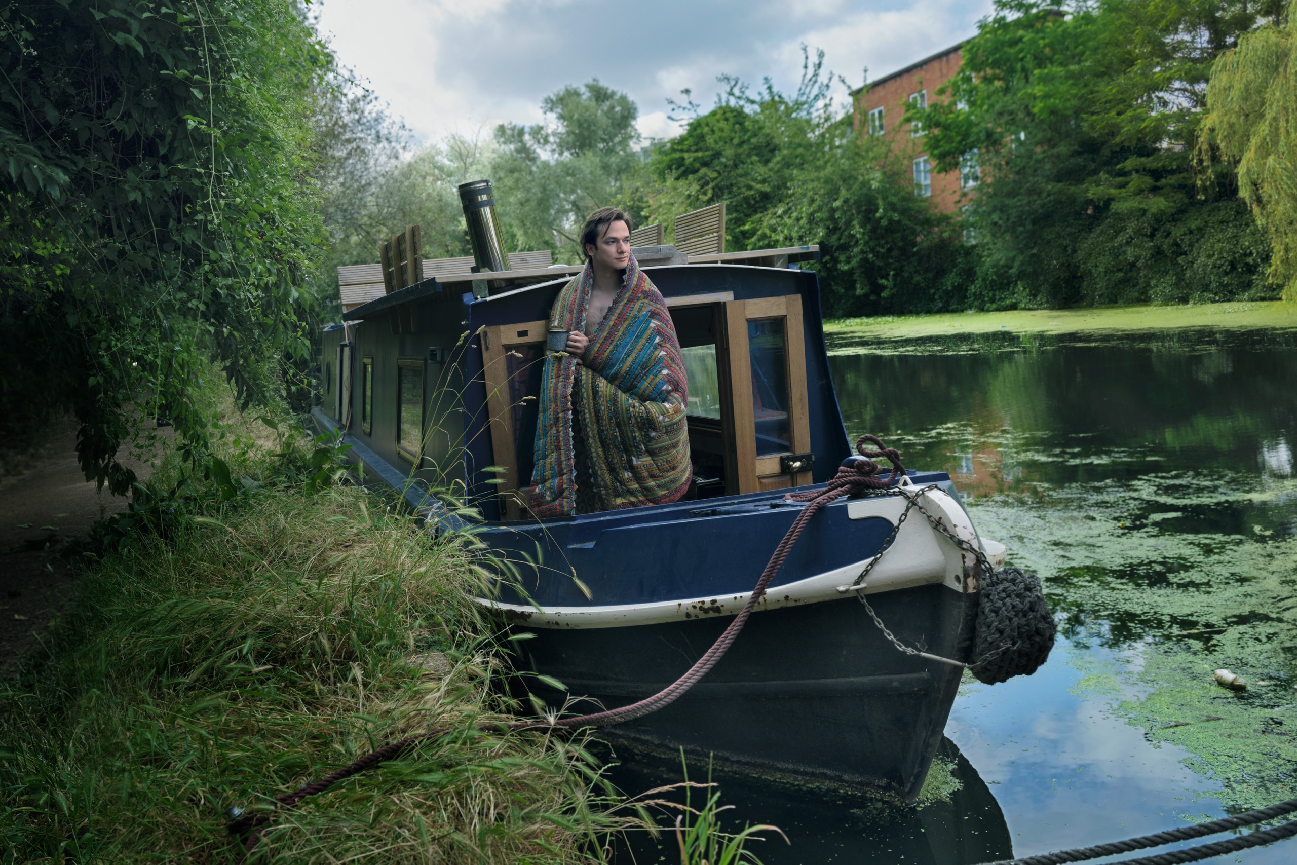 Arslan standing on the boat deck, a blanket coervs his shoulders while he looks out over the river.