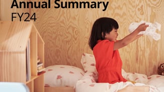 A child wearing a red dress sits on a bed playing with the soft toy SKOGSDUVA while smiling big.