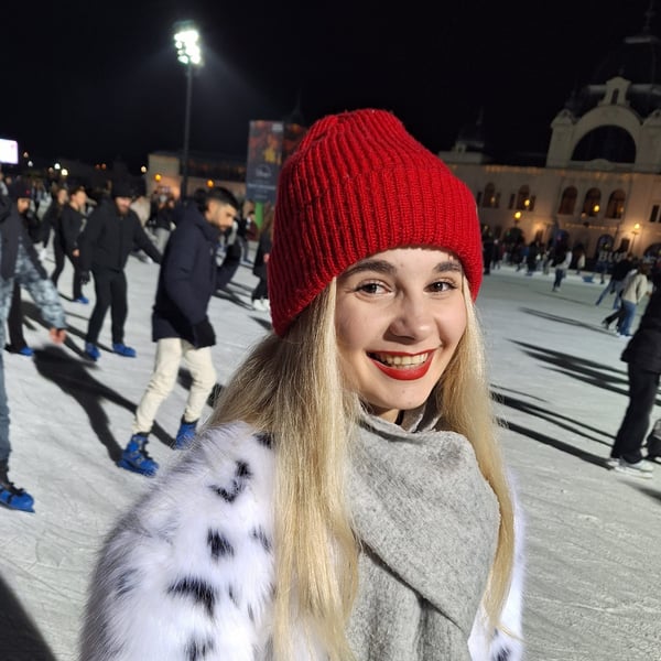 Yana in a red knitted cap smiling on an ice rink with people skating in the background.