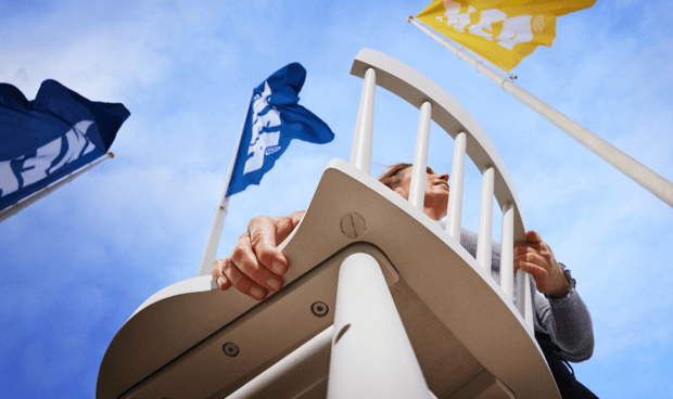 Woman carrying a chair with blue and yellow IKEA flags in the sky behind.