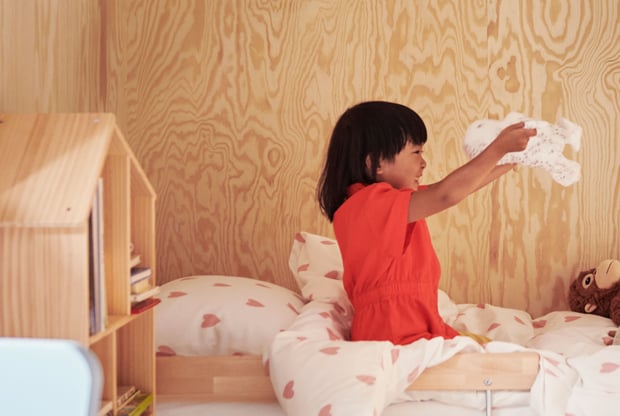 A child wearing a red dress sits on a bed playing with the soft toy SKOGSDUVA while smiling big.