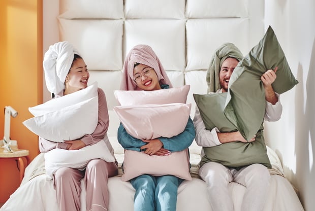 Three people in pyjamas sitting on a bed holding fluffy pillows and laughing.