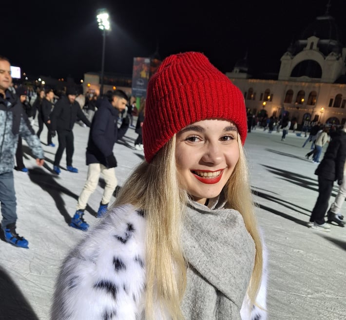 Yana in a red knitted cap smiling on an ice rink with people skating in the background.