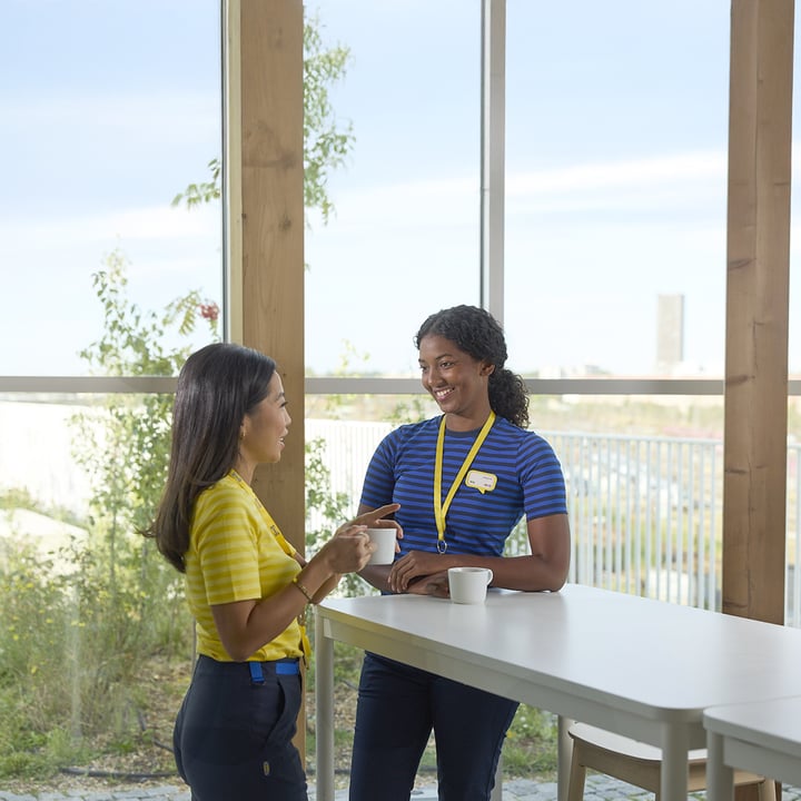 Two IKEA employees talking and having coffee at a tall table in a bright room with large windows and a city view.