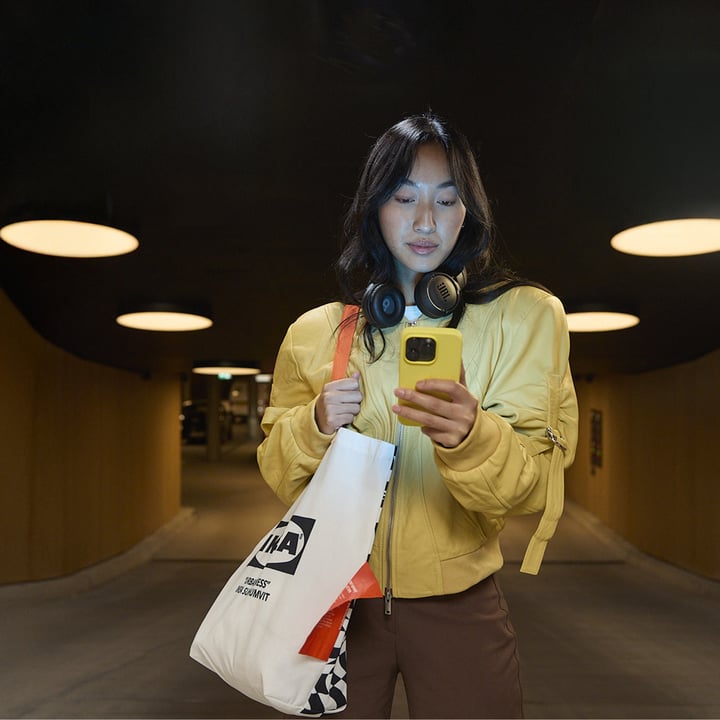 Person in a yellow jacket holding a phone and an IKEA shopping bag, standing in a modern hallway with circular ceiling lights.