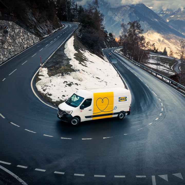 IKEA delivery van driving on a winding mountain road with snow patches and scenic valley views in the background.