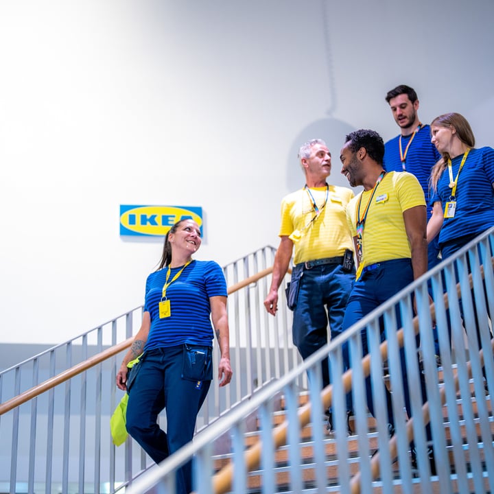 A group of IKEA employees in blue and yellow uniforms walking down a staircase inside an IKEA store, with an IKEA logo visible on the wall.