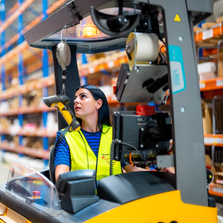 IKEA warehouse worker in a yellow safety vest operating a forklift in a large storage facility with shelves filled with products.
