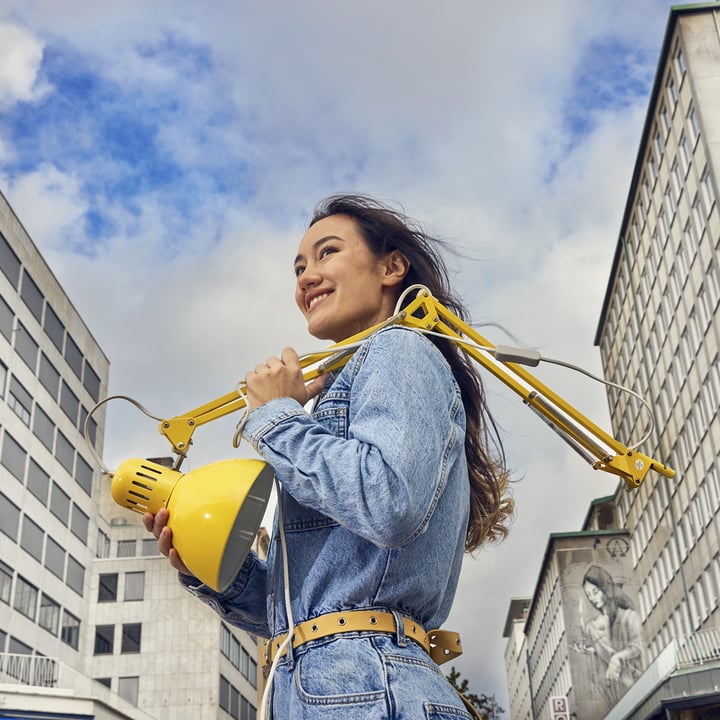 Person in a denim jacket carrying a large yellow desk lamp over their shoulder, standing between tall urban buildings under a partly cloudy sky.