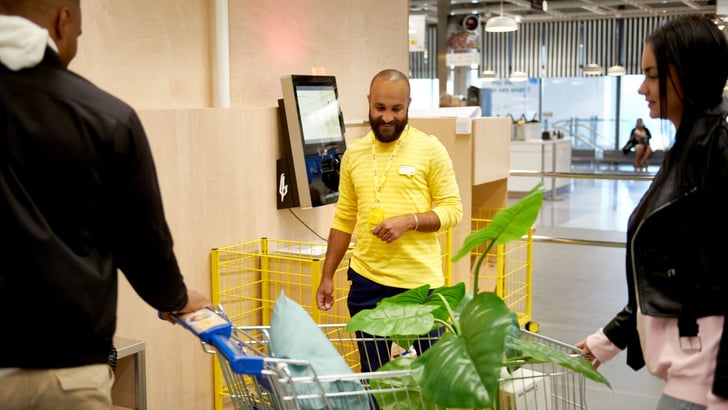 A smiling IKEA employee helping two customers in the self service check-out area.