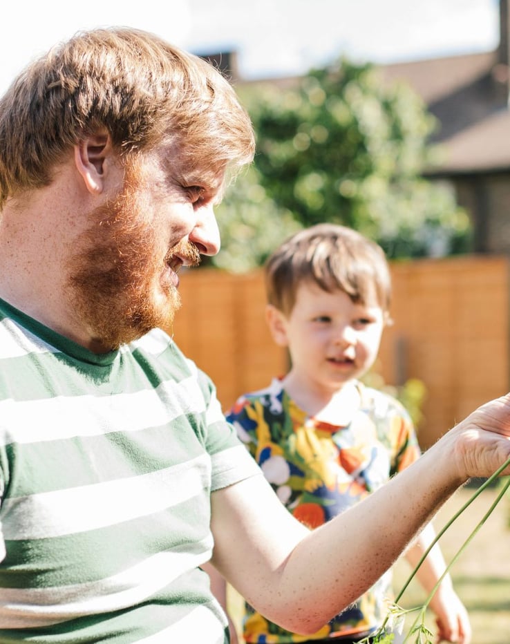 A father holding a homegrown carrot, his small child stands besides him.