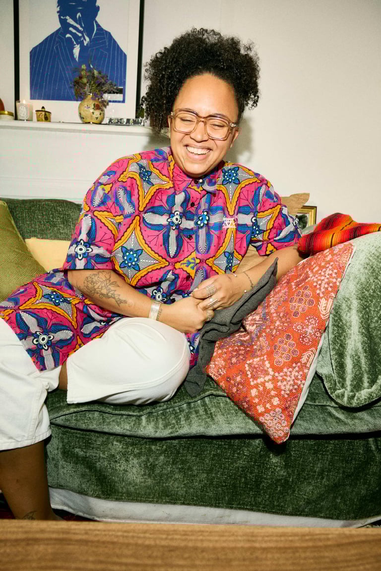 A woman in glasses and a colorful patterned shirt sits on a couch, laughing.