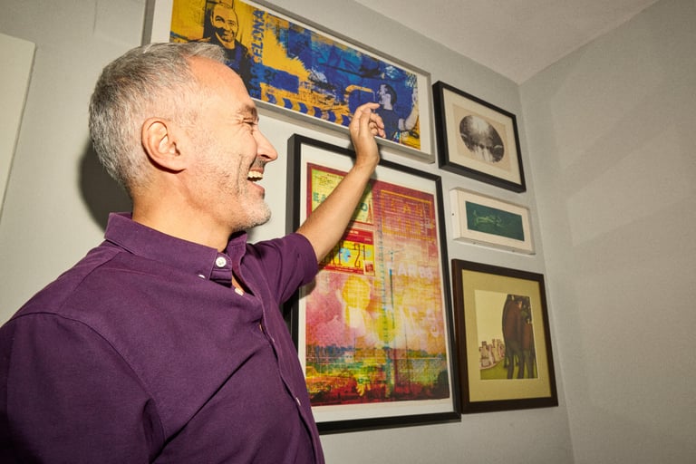 A man smiling and pointing to framed art on a wall.