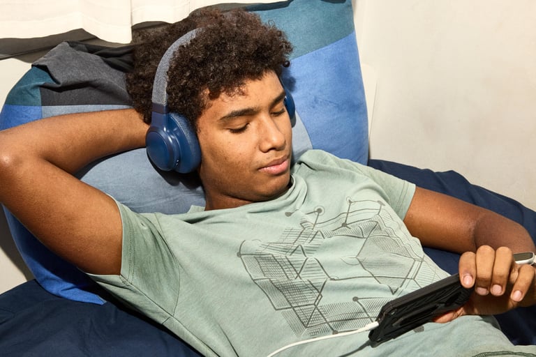A young man with headphones lying on a bed, looking at a smartphone.