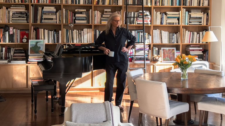 Annie Leibovitz stands beside a piano in front of a bookshelf.