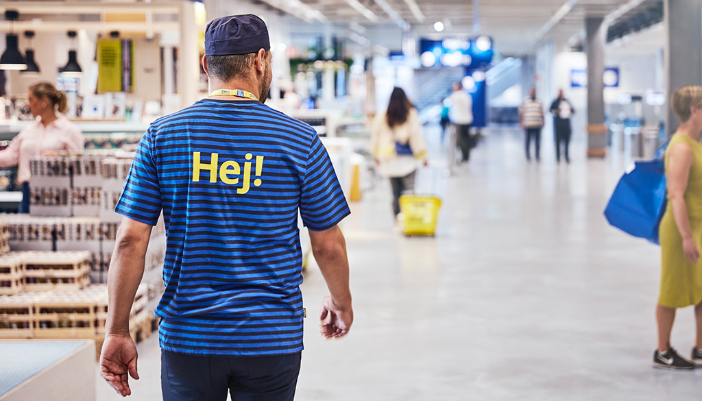The back of a male IKEA employee walking through the store wearing a blue striped t-shirt with Hej! typed in yellow on the back.